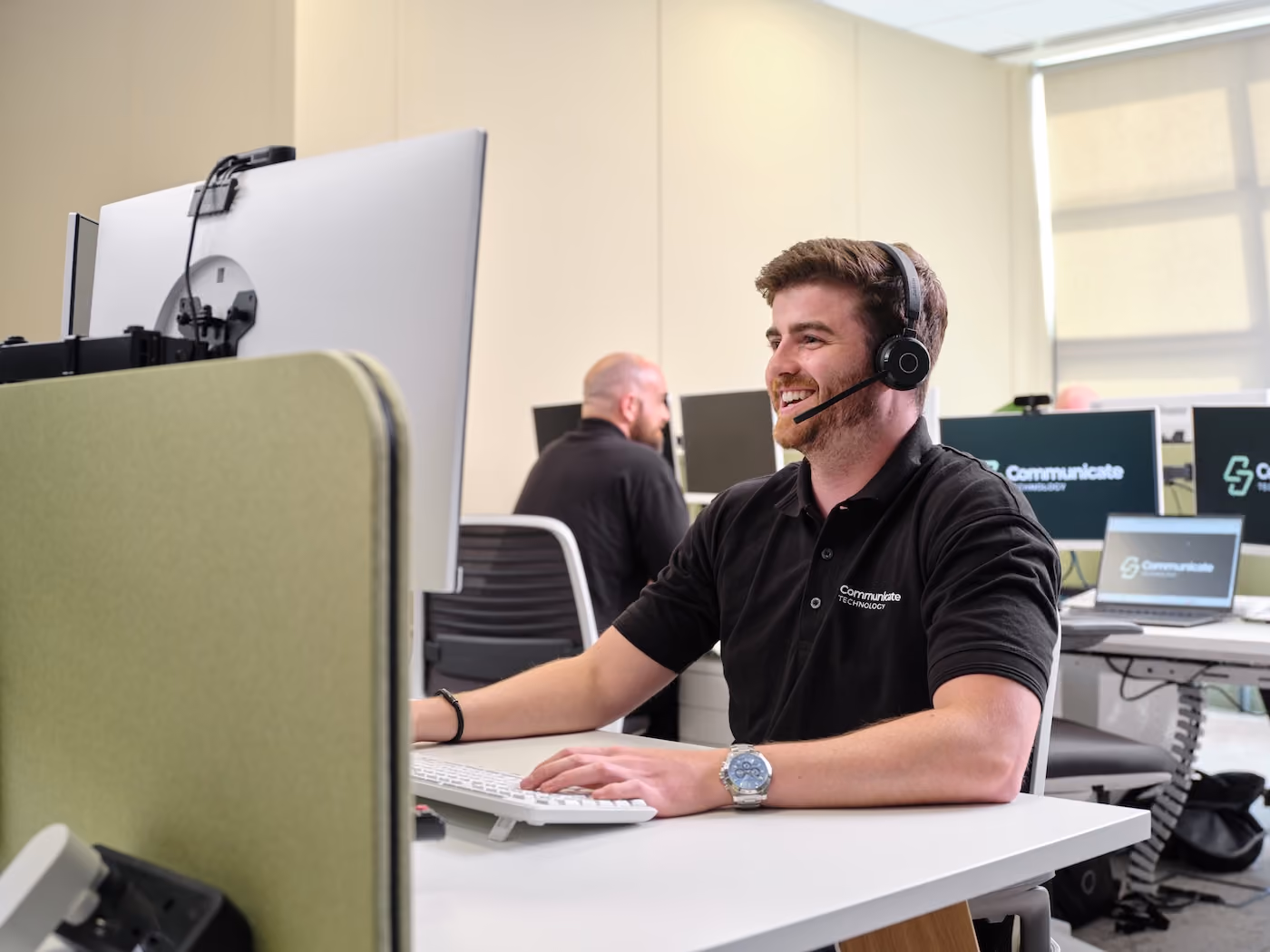 Man with headset smiling while working on a computer in a modern office space with others.