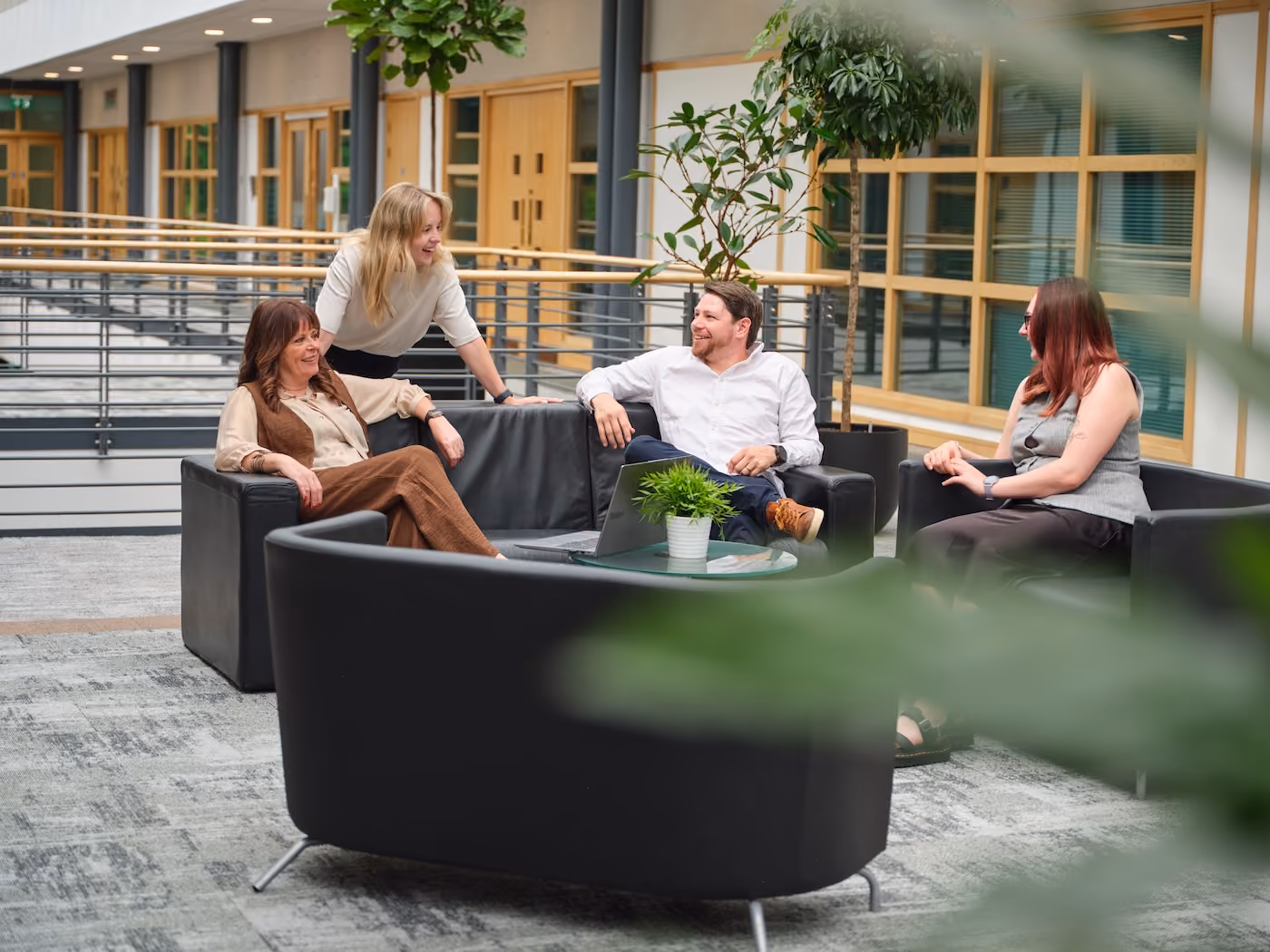 Four people chatting and smiling in a modern office lounge with black sofas and green plants.