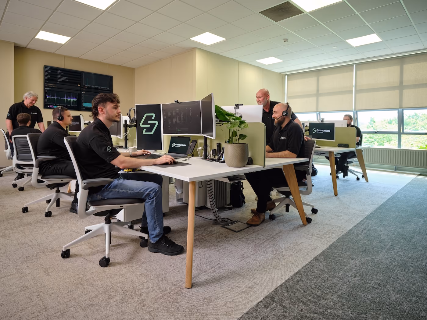 Office with multiple employees working at desks with computers, some wearing headsets, in a bright workspace.