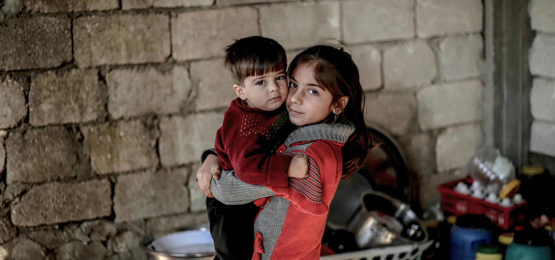 oung girl holding her little brother in front of a stone wall, illustrating Karuna's story on rebuilding childhood after conflict