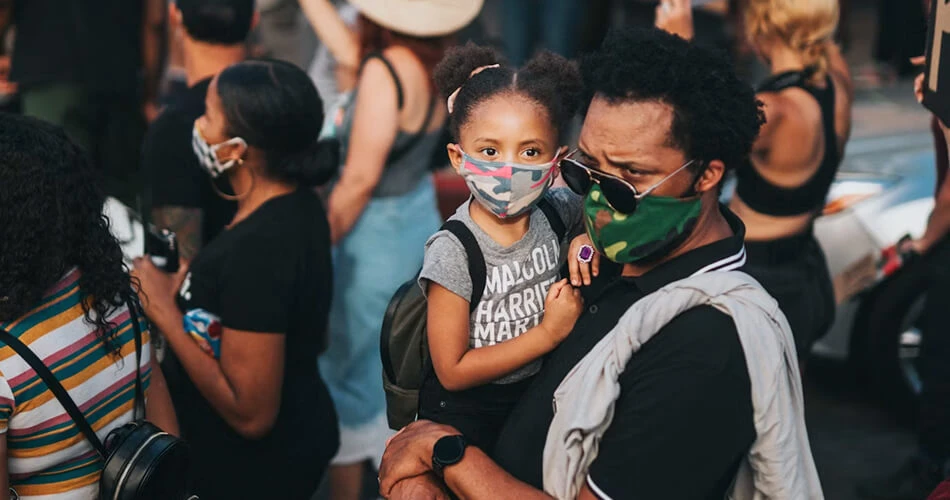 Alt text: "Father carrying young daughter wearing protective masks in a crowd at a community gathering