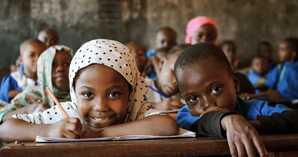 Smiling schoolgirl in a polka dot headscarf writing at a classroom desk, surrounded by classmates in Africa