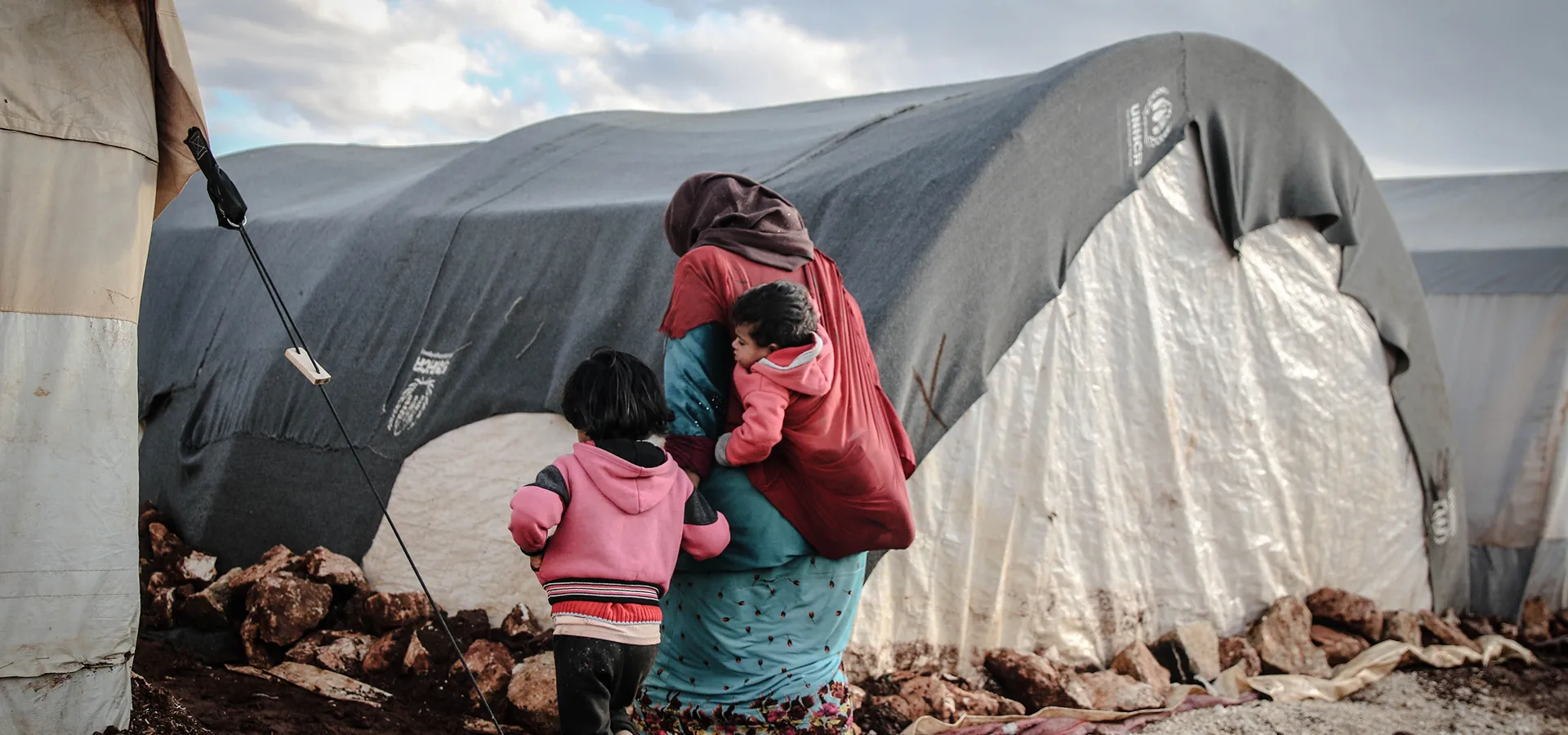 Mother carrying young children beside a tent at a refugee camp