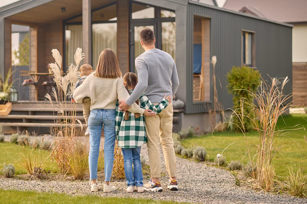 Family of four standing arm in arm facing modern house with gray exterior and wooden porch.