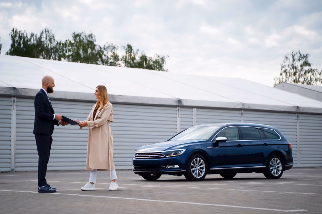 Man handing car keys and documents to woman next to a blue station wagon in a parking lot.