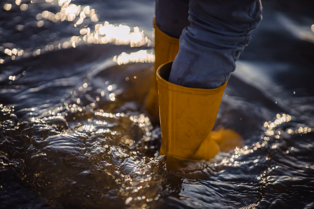 Person wearing yellow boots walking through shallow water with sunlight reflecting on the surface.