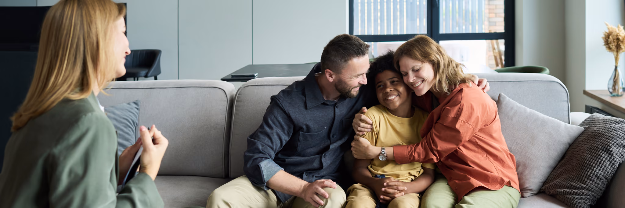 Smiling parents hug their child on a couch while talking with a woman, possibly a therapist, at home.