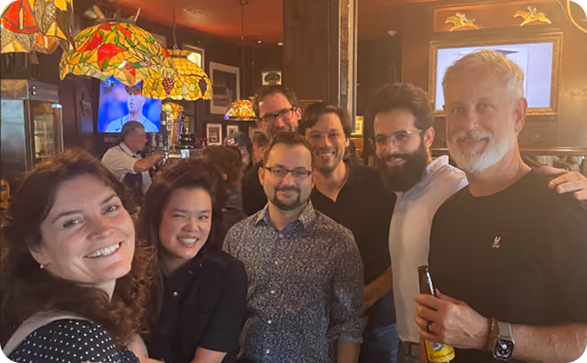 Group of seven adults smiling and posing together in a warmly lit bar or pub setting.