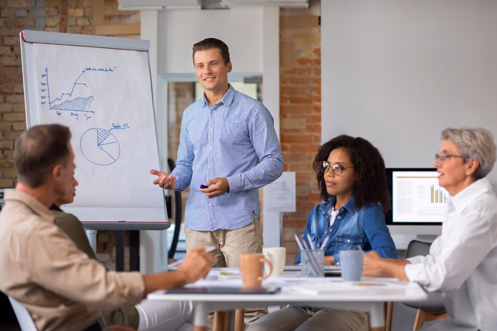 Man in blue shirt presenting charts on flip chart to three colleagues in office meeting.