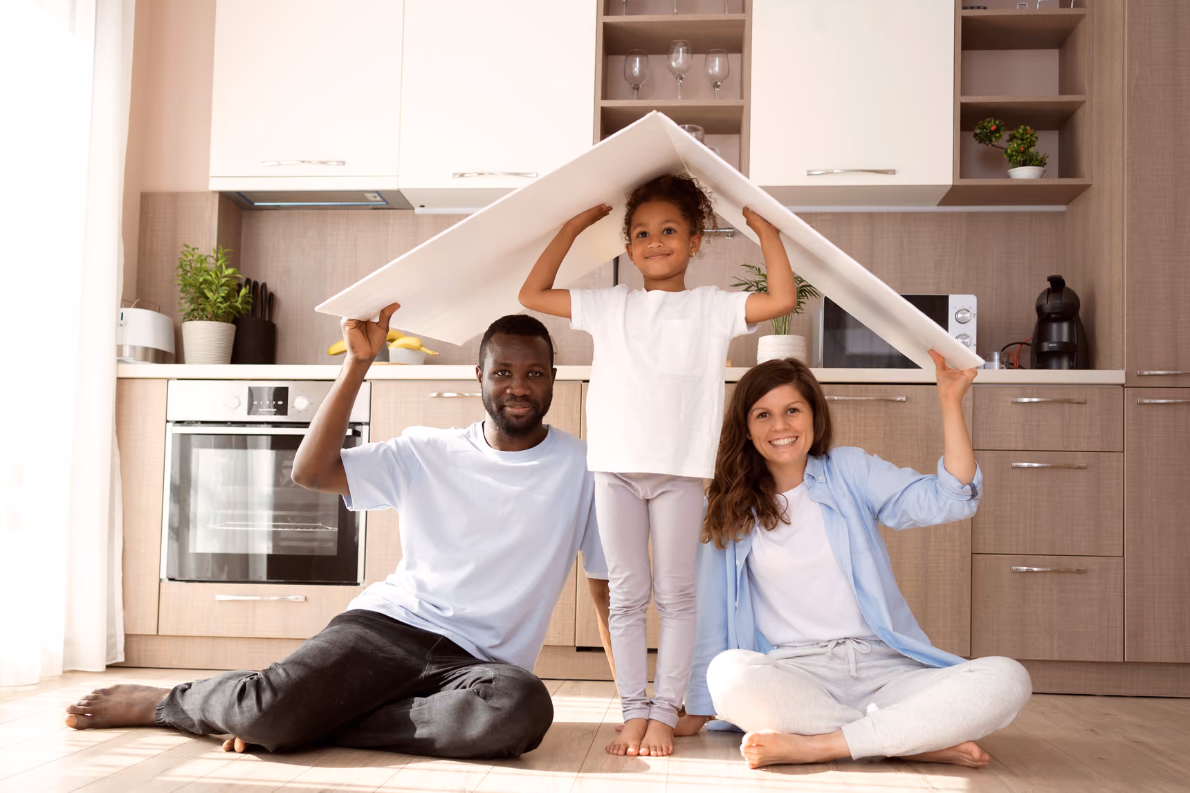 Smiling family of three sitting on a kitchen floor under a makeshift roof made from a large white board.