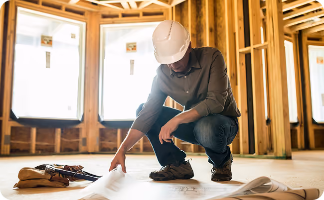 Construction worker wearing a hard hat kneeling on floor reviewing blueprints inside a framed building.