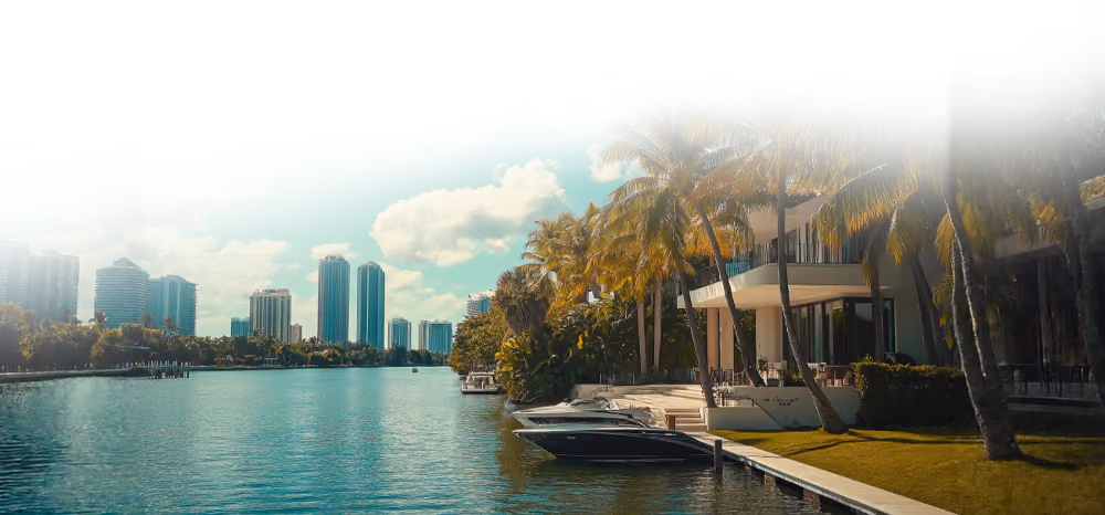 Waterfront view with boats, palm trees, modern buildings, and partly cloudy blue sky.
