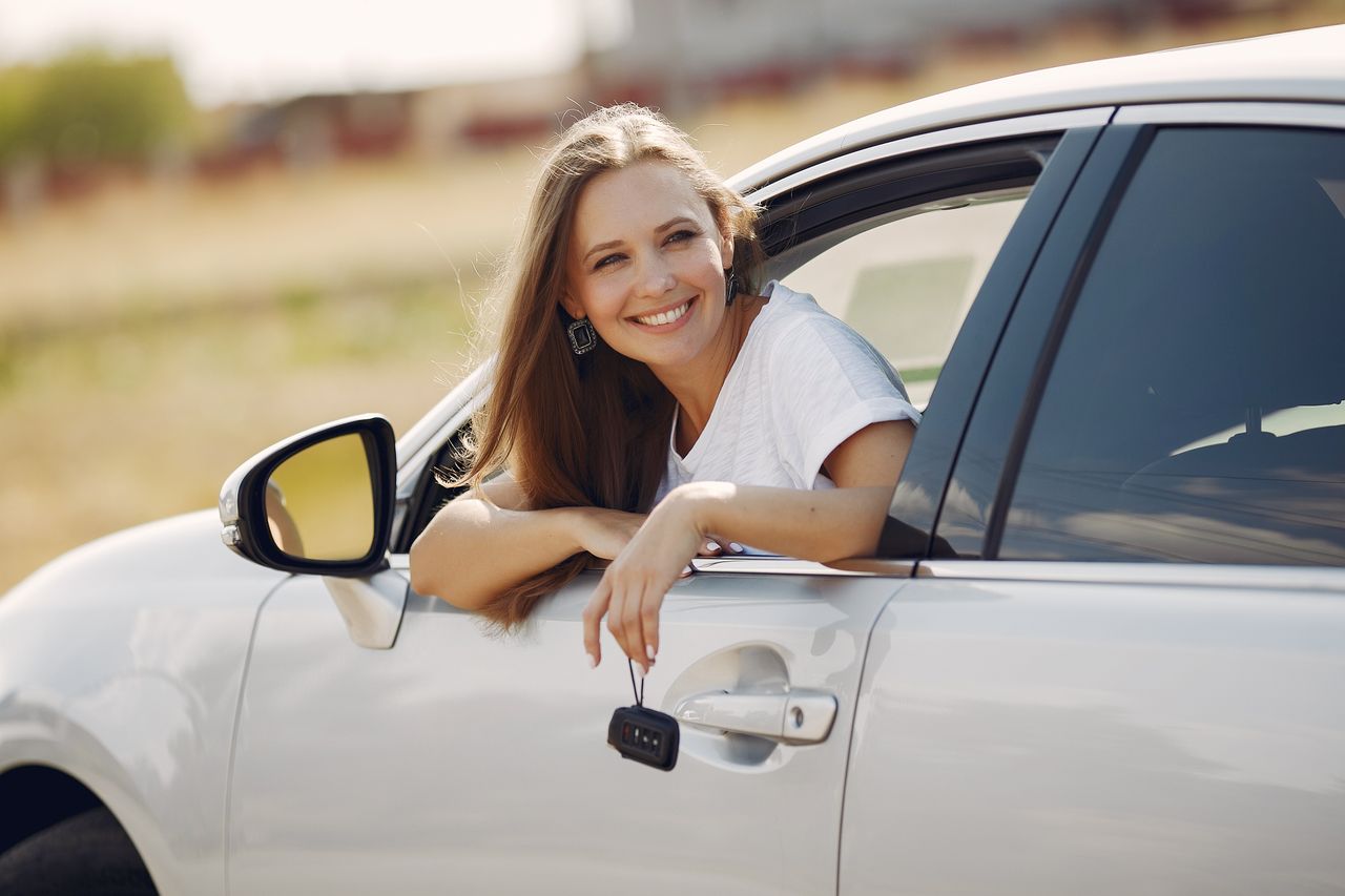 Smiling woman leaning out of a white car window holding car keys in her hand.