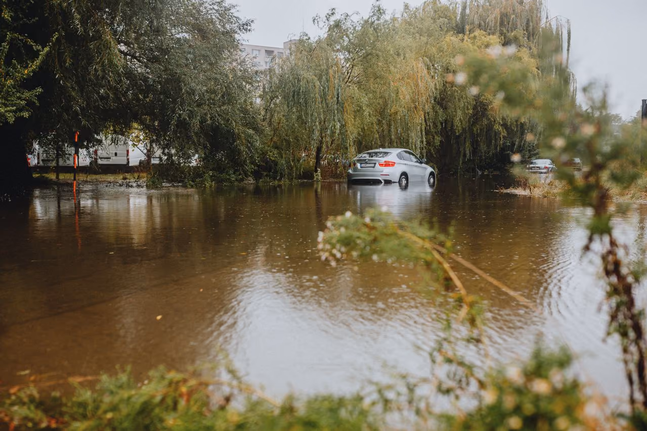 Silver car partially submerged in floodwater on a tree-lined street with a second car in the background.