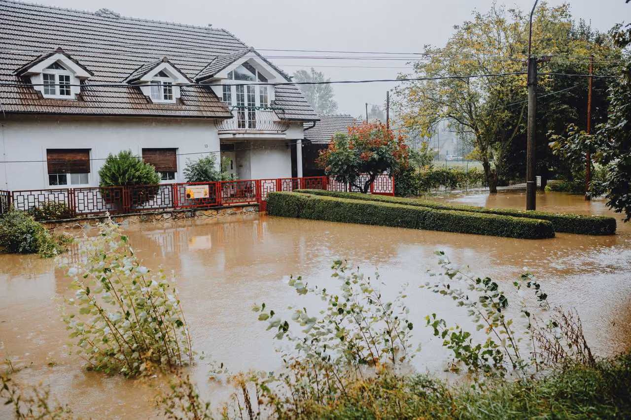 Floodwaters partially submerge yard, bushes, and street outside a two-story house on a rainy day.
