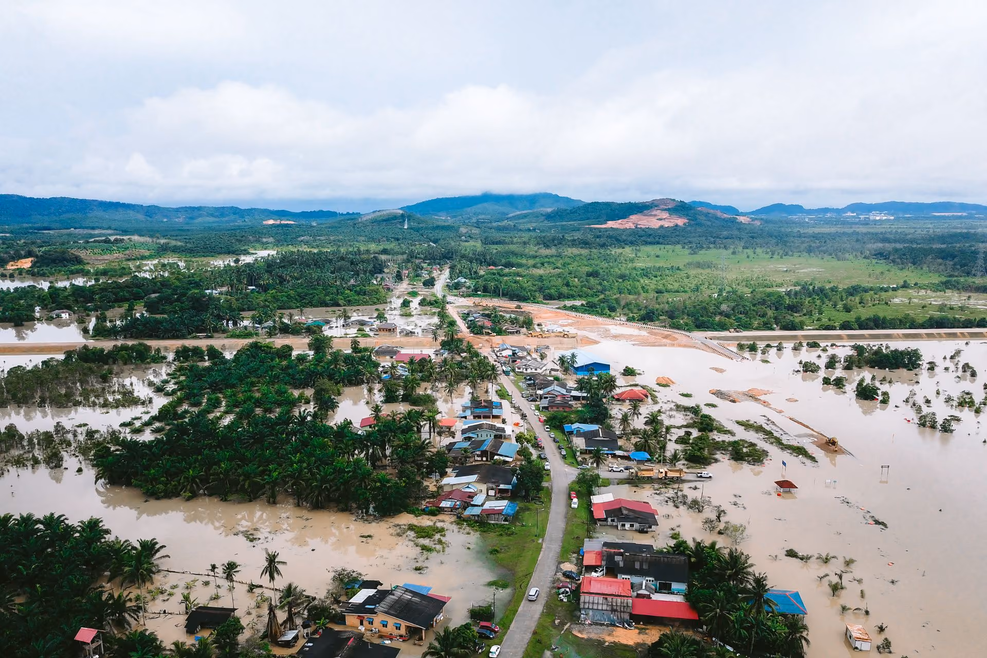 Aerial view of a flooded village with many houses surrounded by muddy water and green hills in the background.