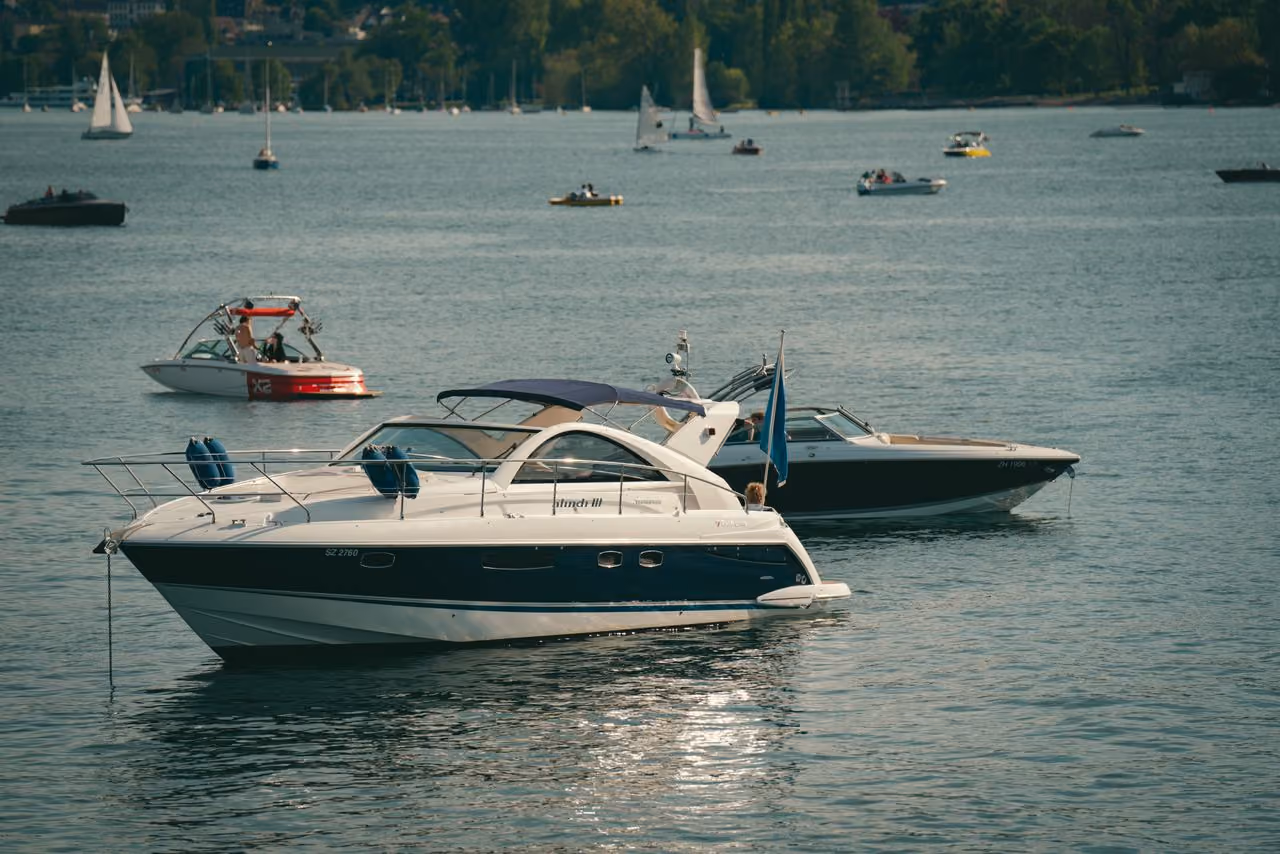 Two motorboats on a lake with people, sailboats, and trees in the distance on a sunny day.