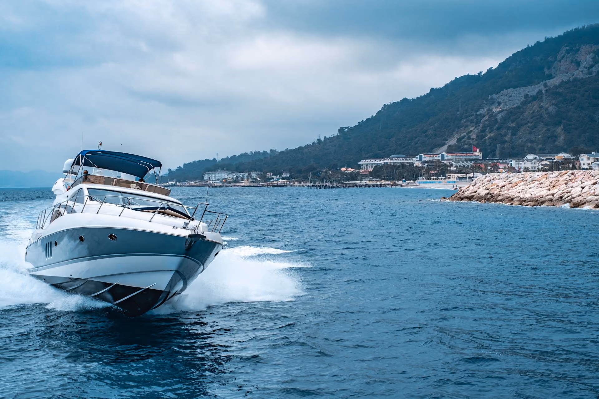 White motor yacht speeding on blue ocean water near a rocky shore and forested hills under cloudy sky.