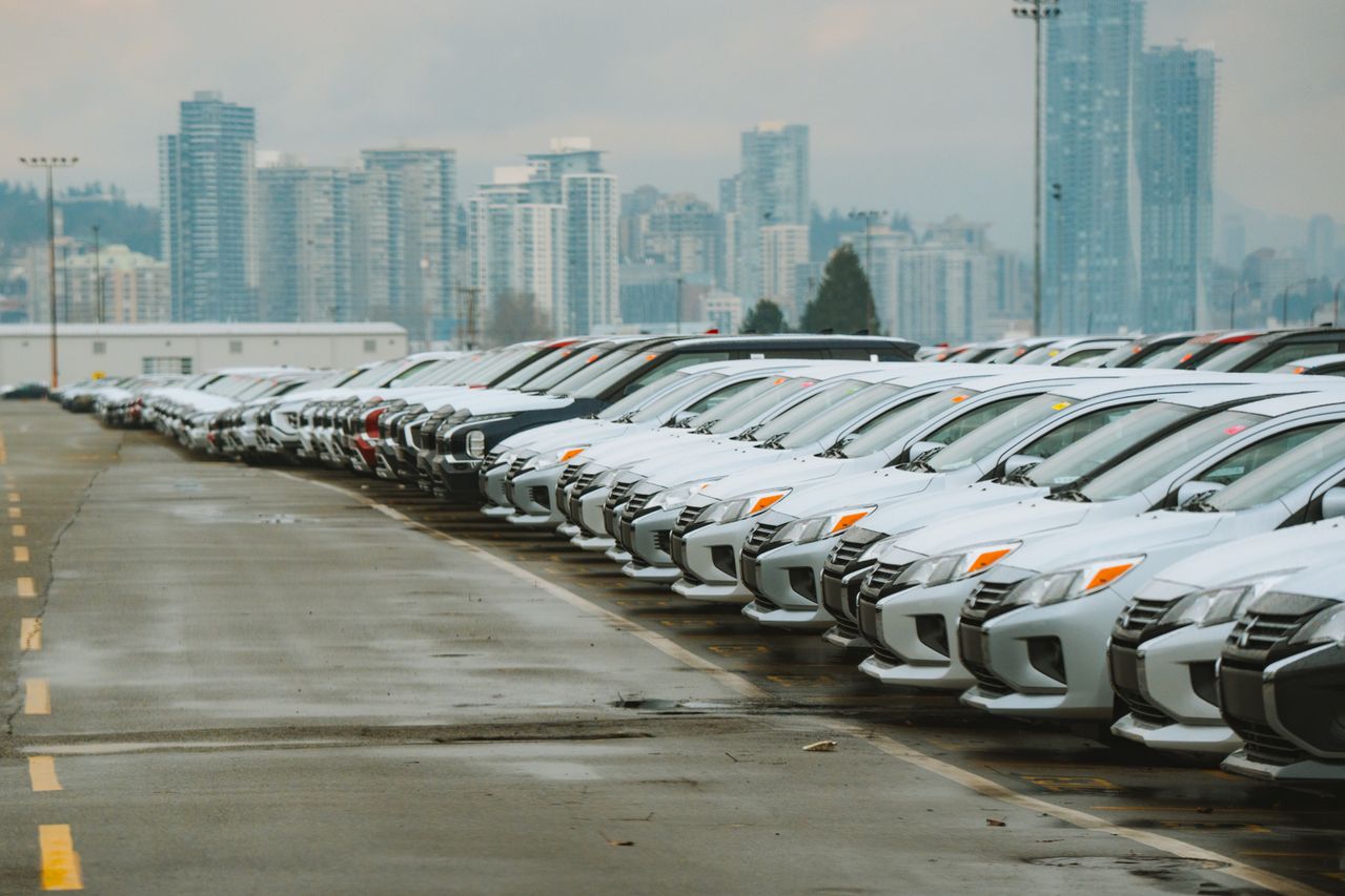 Rows of mostly white cars parked in a large lot with city buildings in the background.