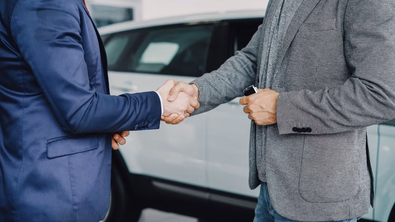 Two men shaking hands in front of a white car, one holding car keys, wearing business casual attire.