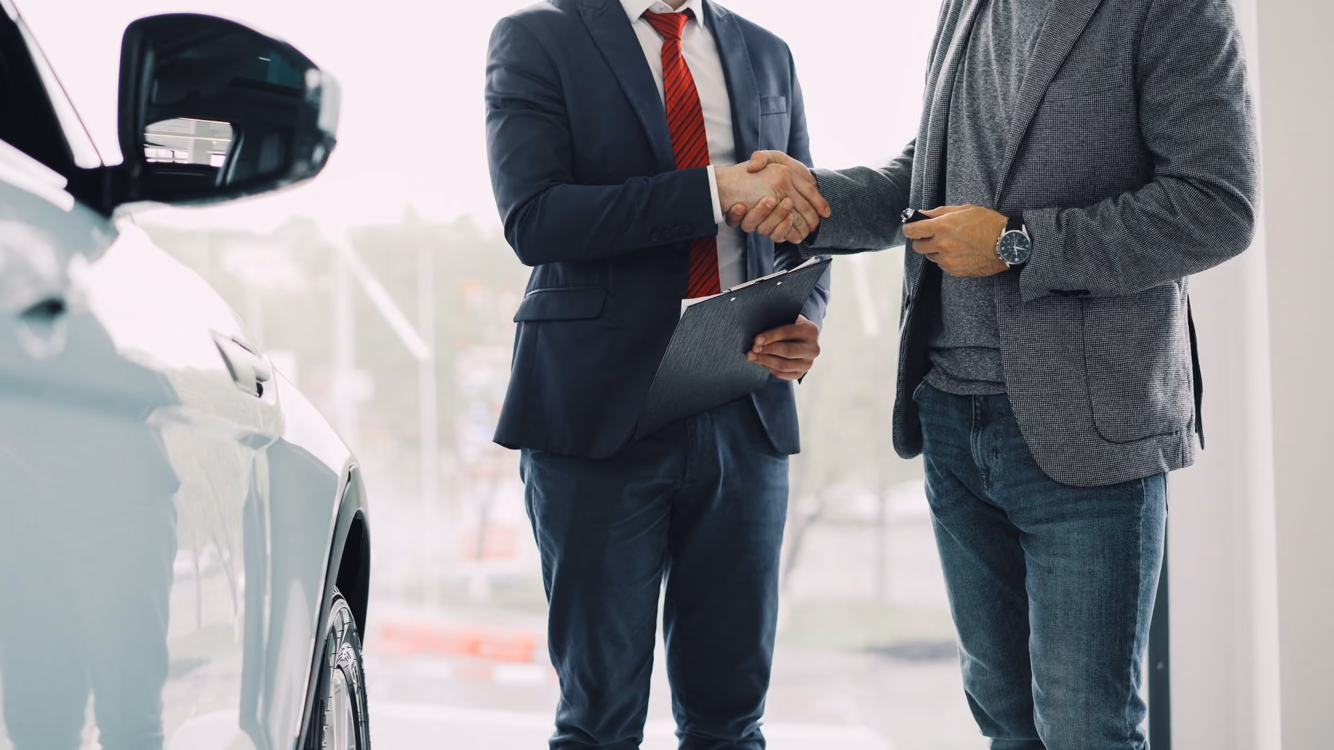 Two men shaking hands next to a white car, one holding a clipboard and the other car keys.