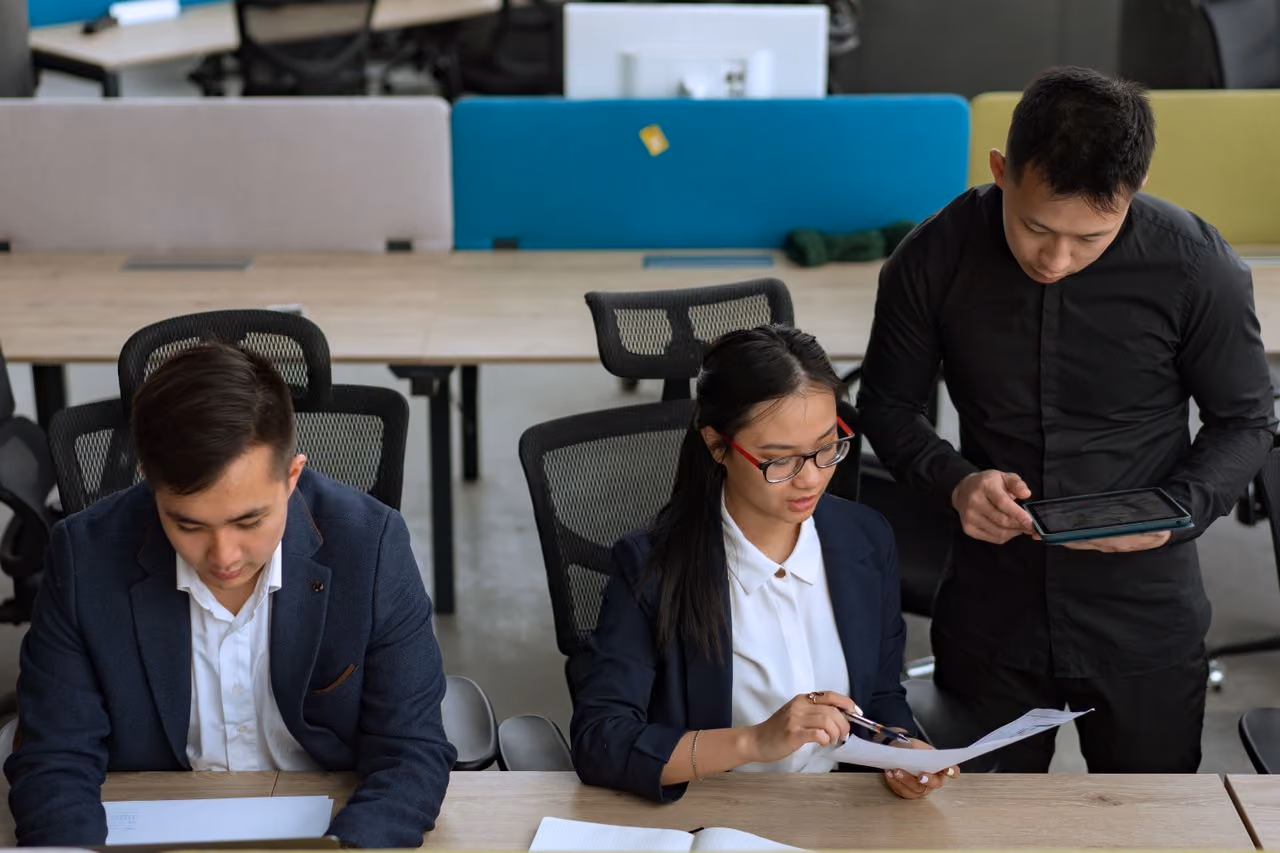 Three professionals in an office reviewing documents and using a tablet at a shared desk.