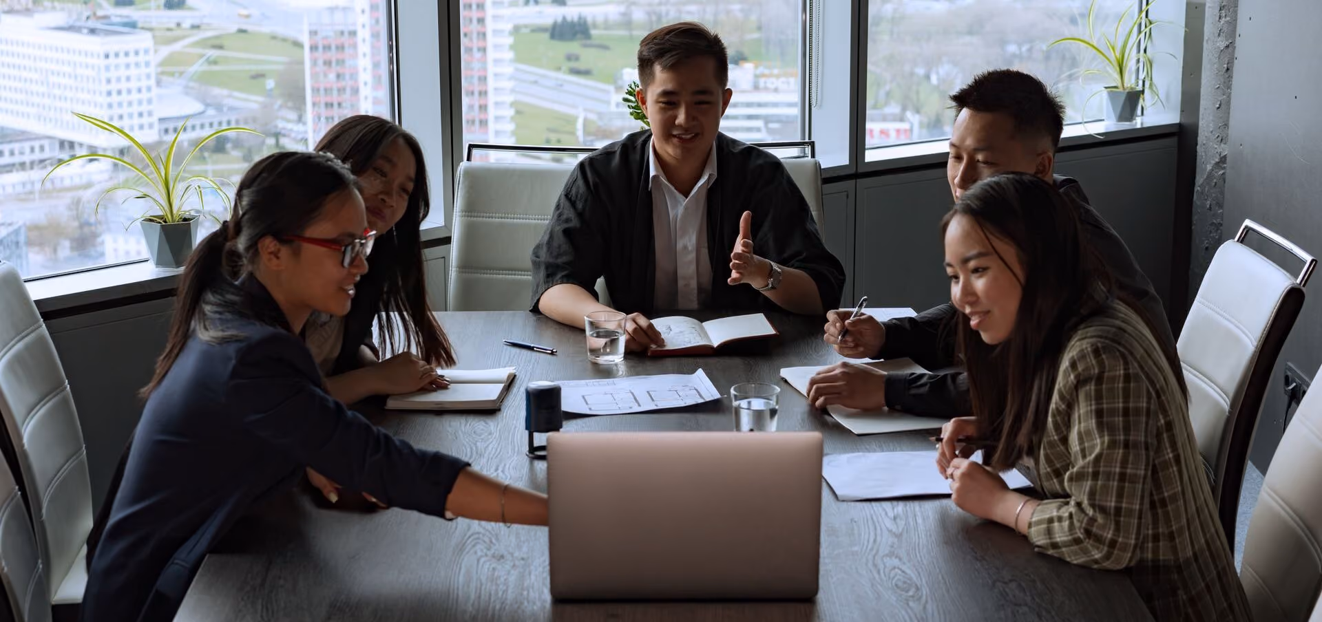 Five young professionals sit at a table, engaged in a meeting with a laptop and notebooks.