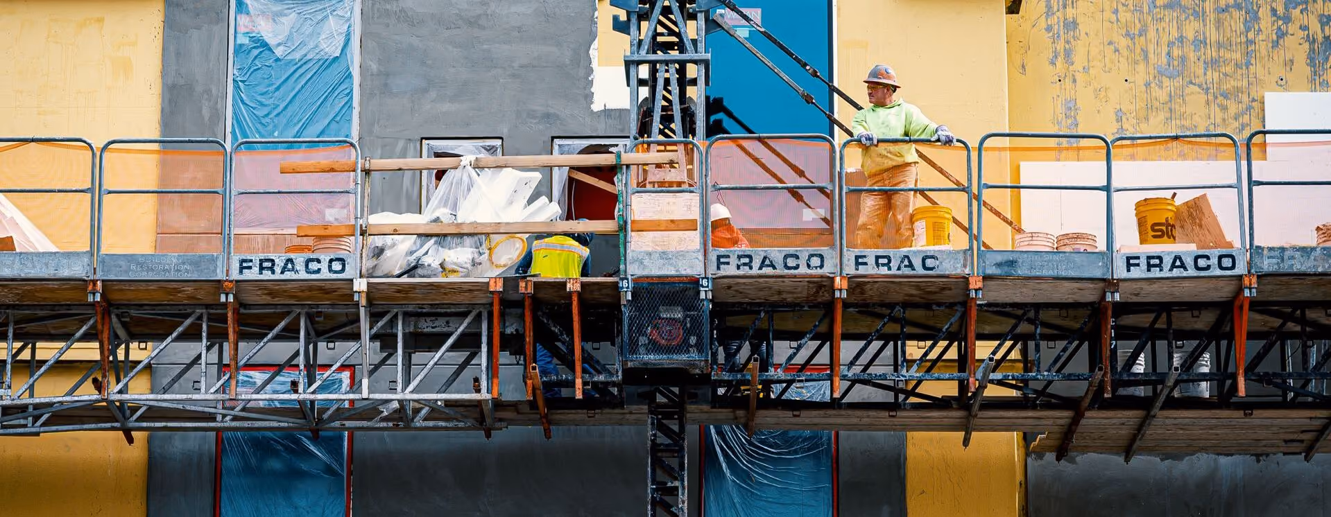 Two construction workers on scaffolding on a building exterior in bright daylight.