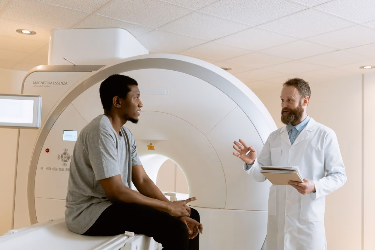 Doctor in white coat talks to seated patient in front of an MRI machine in a medical facility.