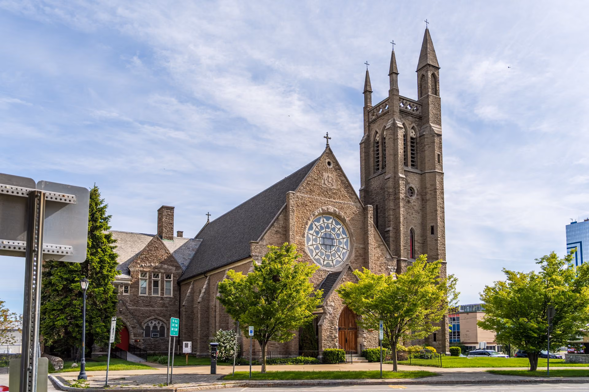 Stone church with tall tower, rose window, wooden doors, and trees in front under a partly cloudy sky.