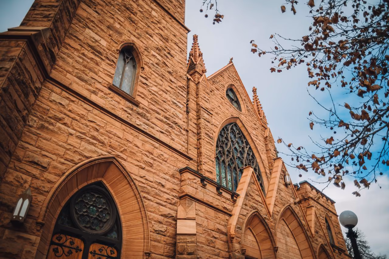 Low angle view of a stone church with arched stained glass windows and a leafless tree branch.