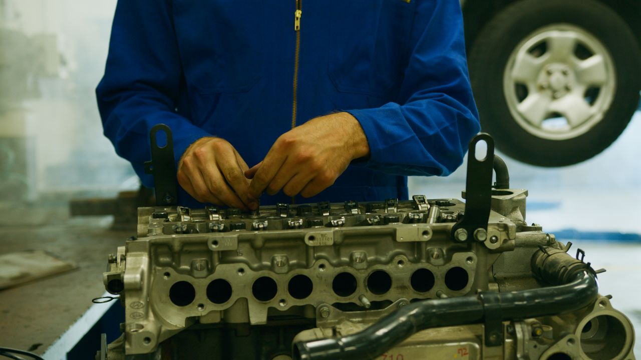 Mechanic in blue coveralls working on a disassembled car engine in a garage.