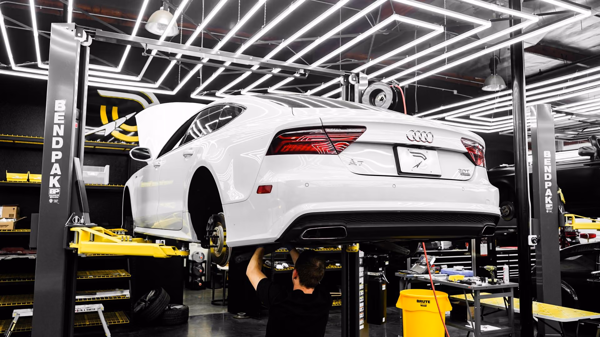 Mechanic working under a white Audi A7 lifted on a BendPak car lift in a brightly lit garage.