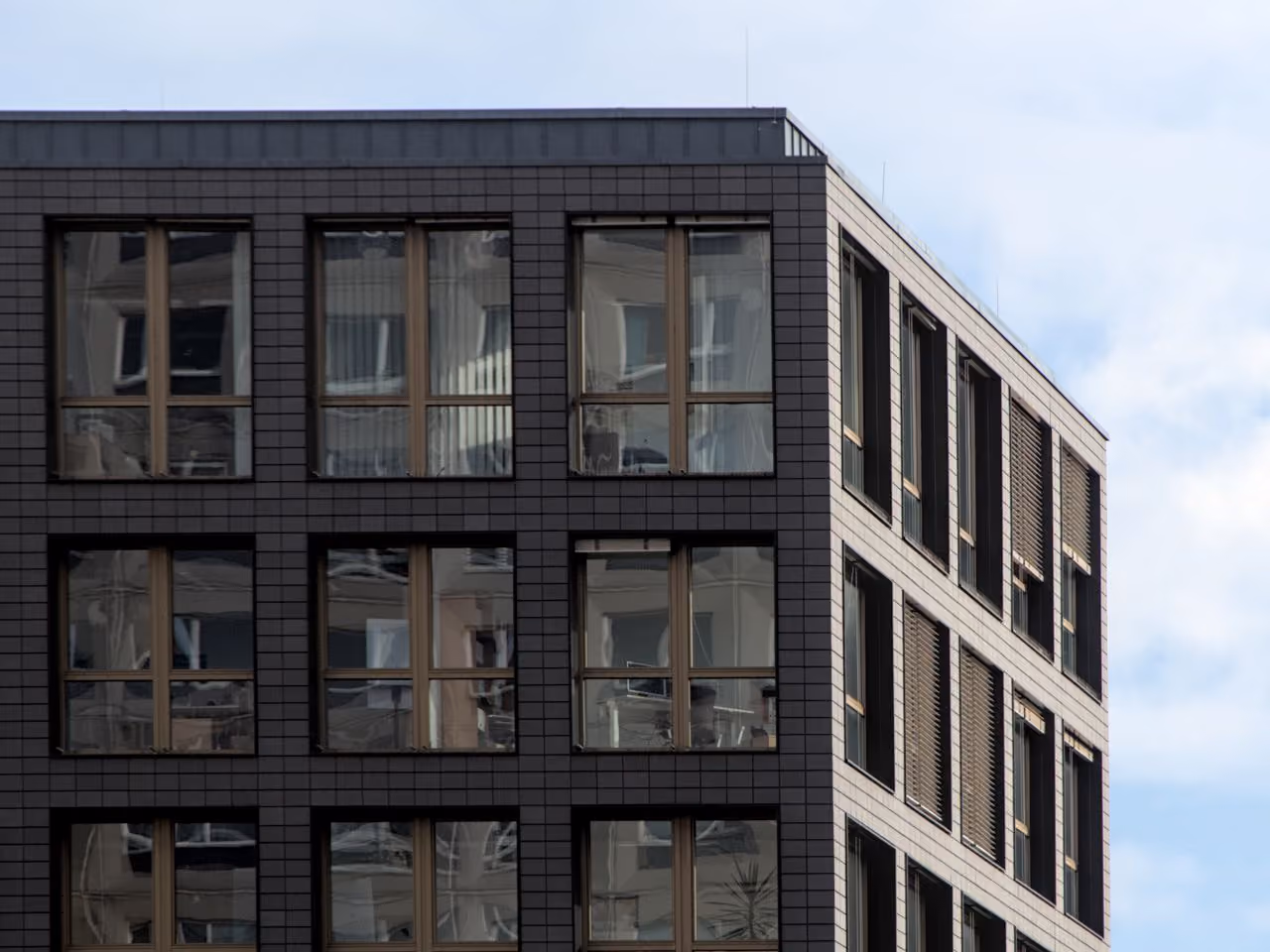 Modern building corner with dark tiled facade and reflective windows under a cloudy sky.
