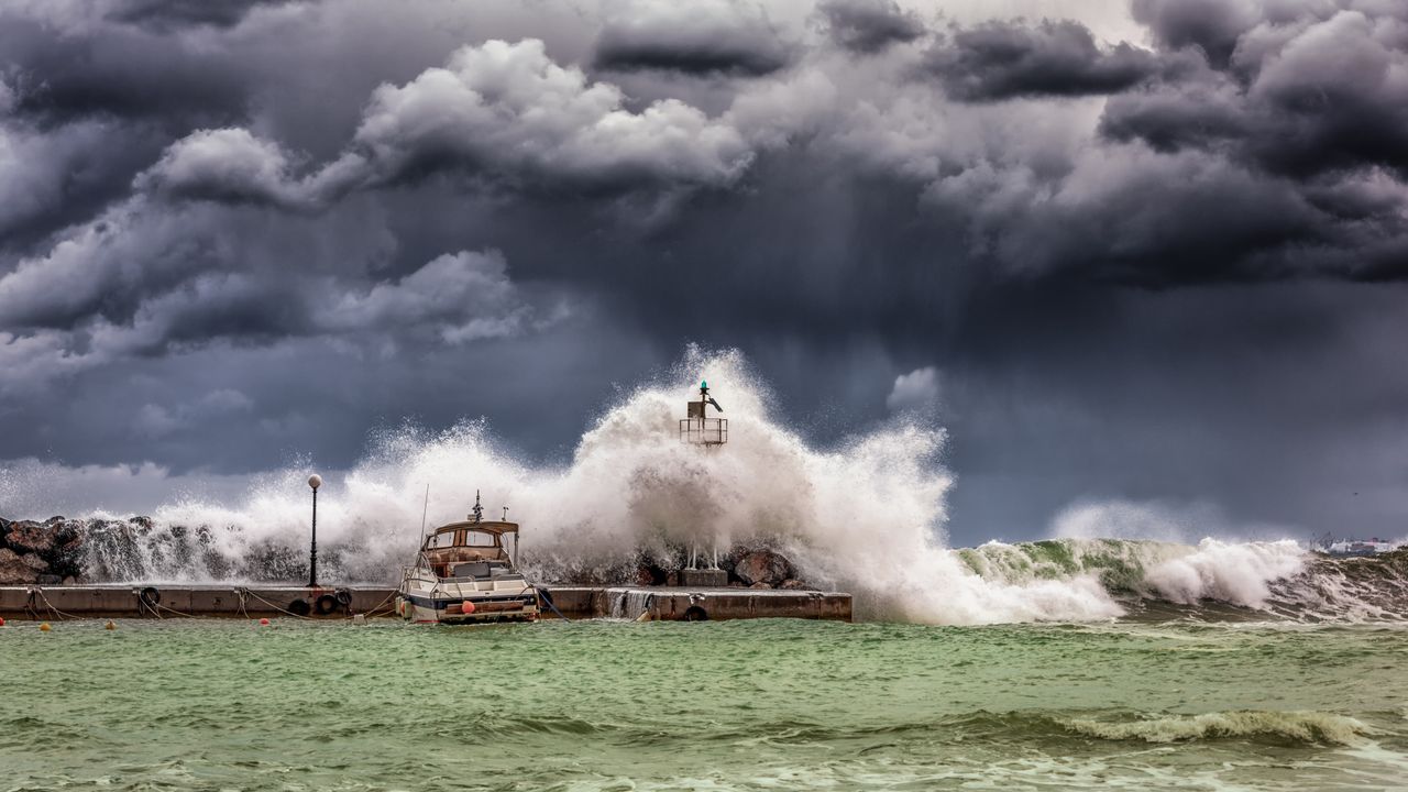 Large waves crashing over a jetty with a boat moored nearby under dark stormy clouds.