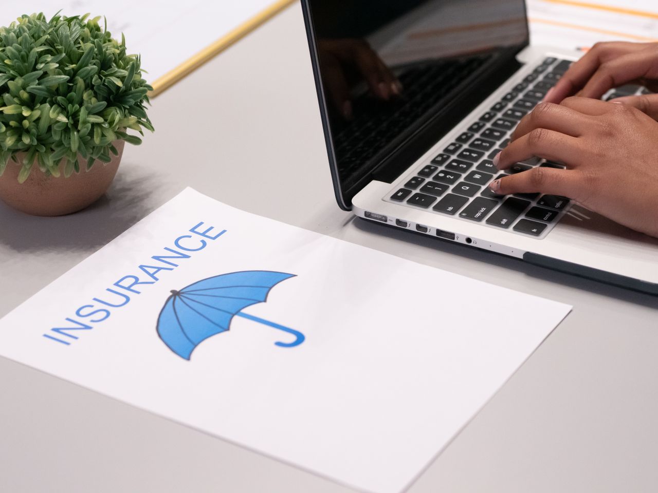 Person typing on a laptop keyboard beside a paper with a blue umbrella and the word INSURANCE.