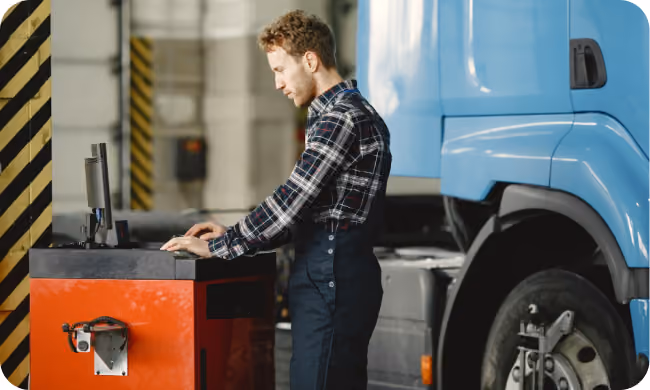 Young man in work overalls using a console in a garage beside a large blue truck.
