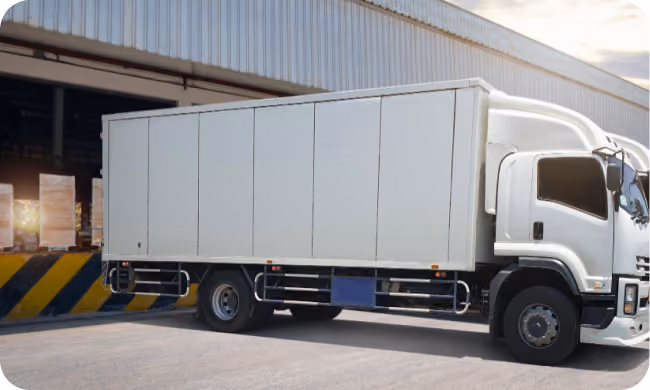 White box truck parked outside a warehouse with loading dock and pallets inside.