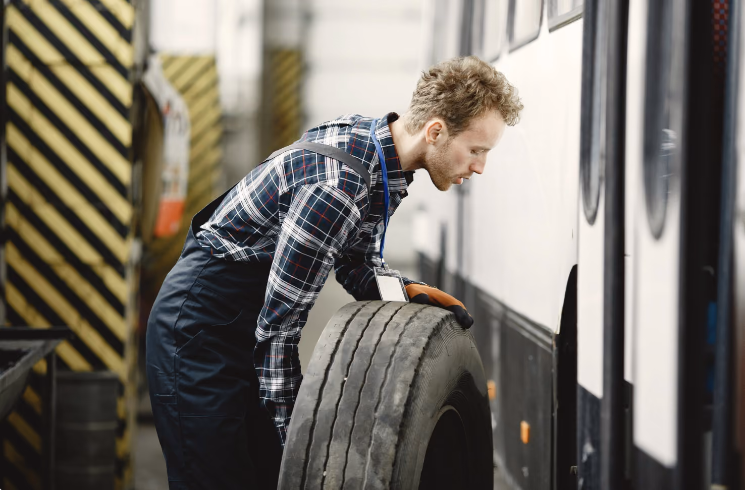 Man in plaid shirt and overalls working on a red machine next to a blue truck in a warehouse.