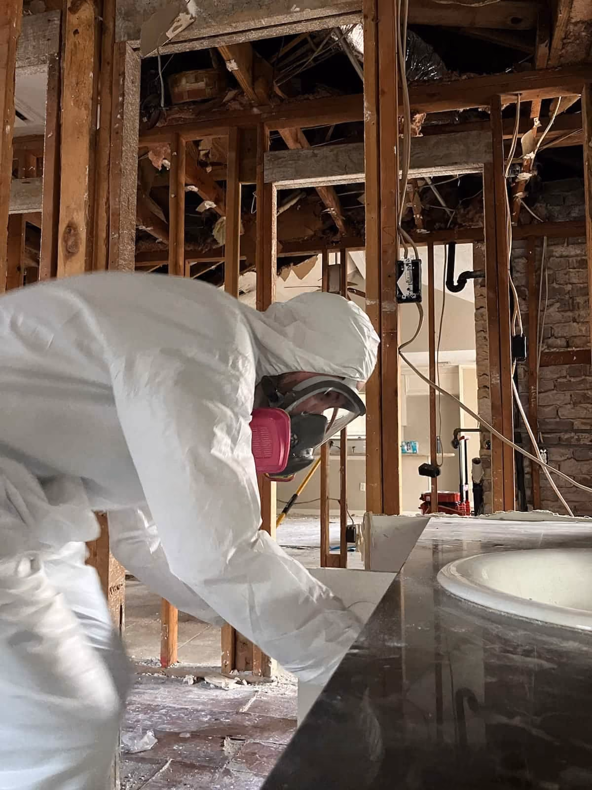 Worker in white protective suit and mask inspecting debris in partially demolished room.