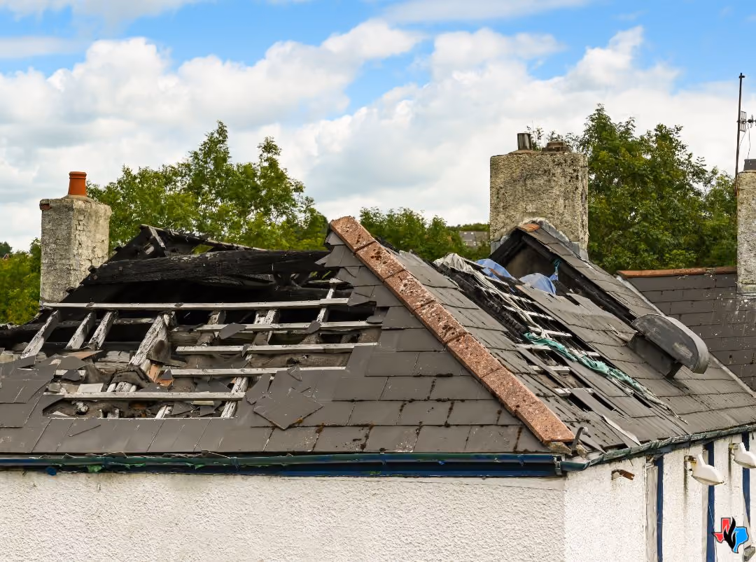 House roof severely damaged with missing tiles and exposed burned wooden beams under blue sky.