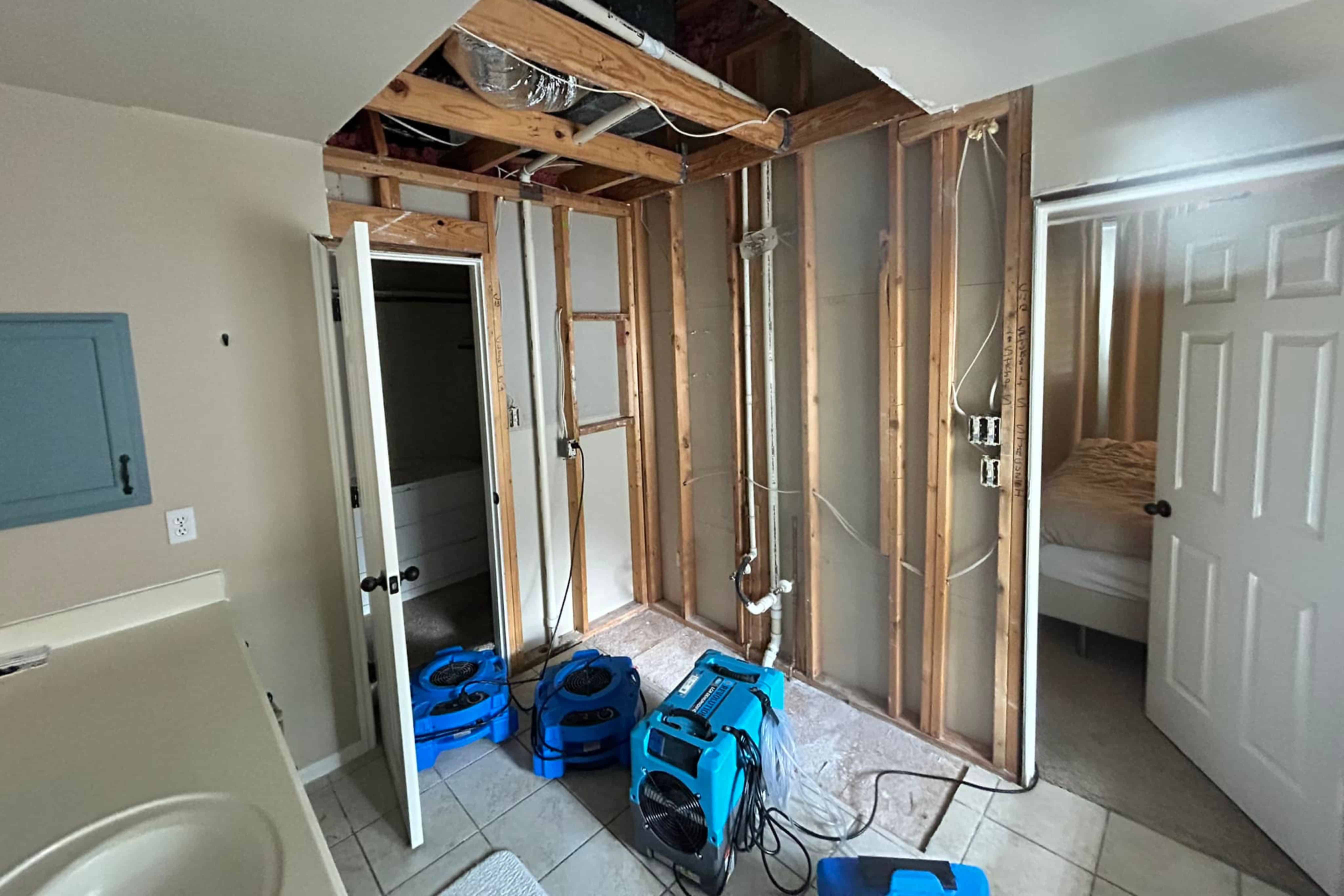 Room under renovation with exposed wood framing, blue air blowers, and open doors to bathroom and bedroom.