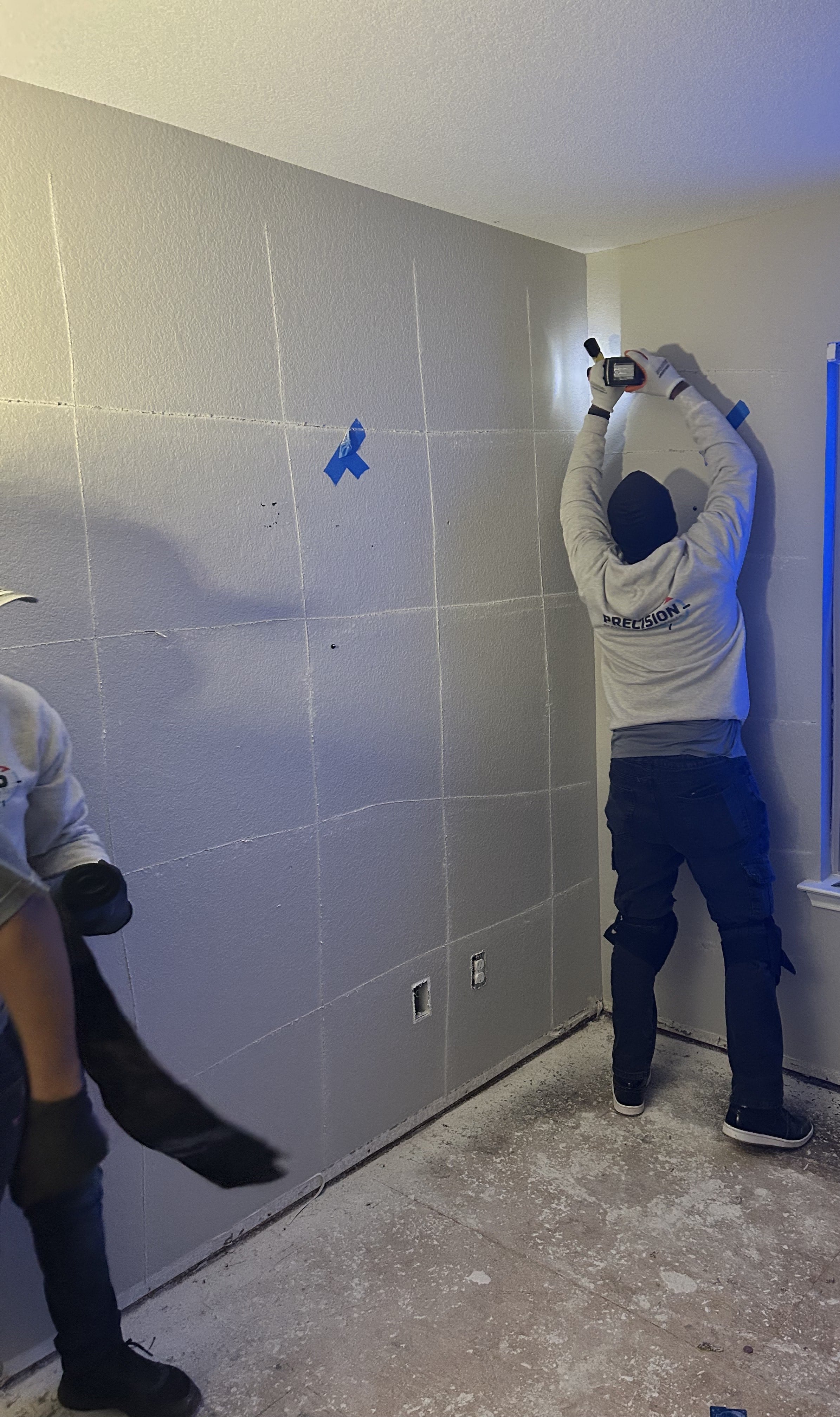 Worker in kneepads using a tool on a wall marked with grid lines and blue painter's tape.