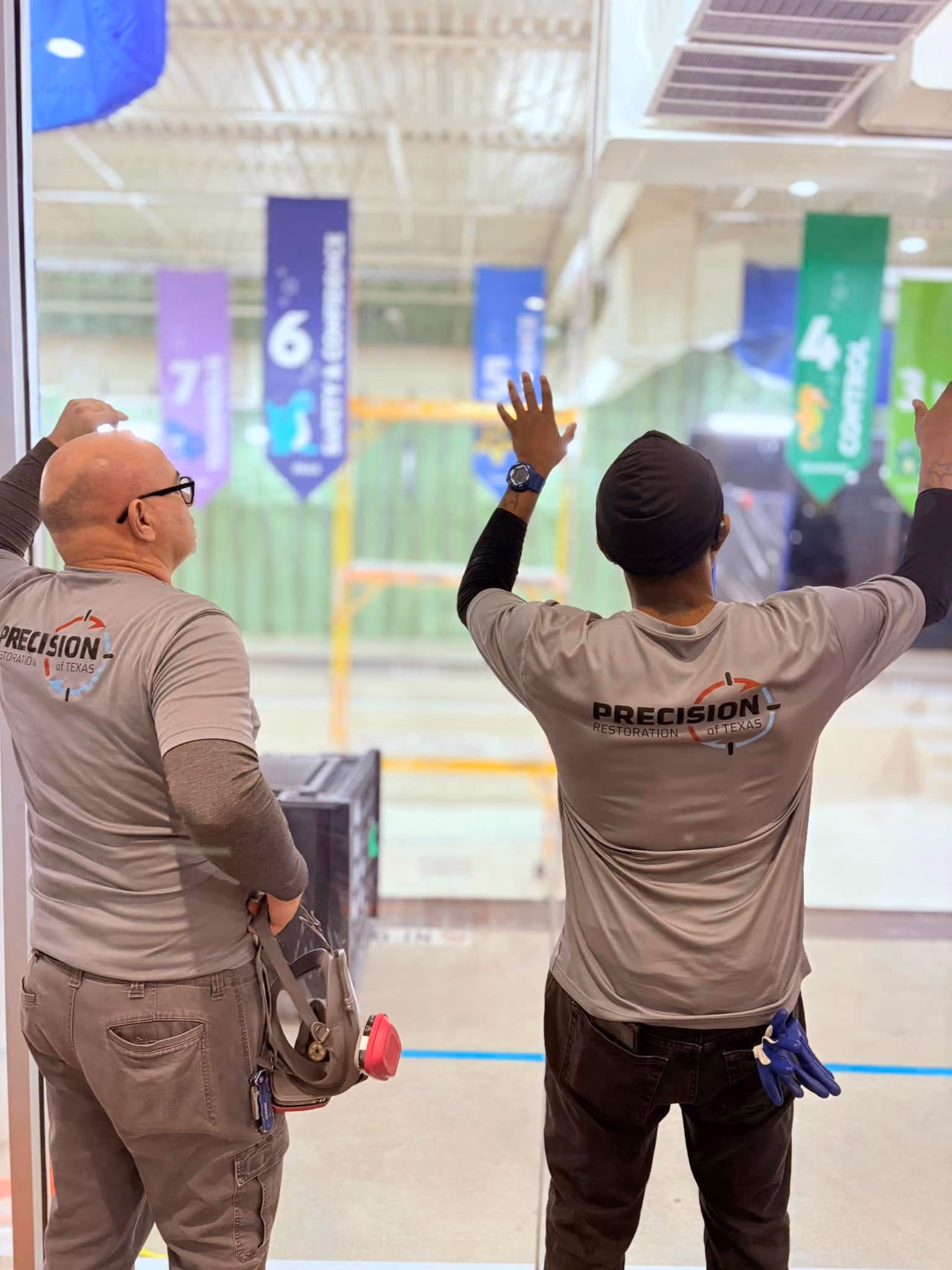 Two men in Precision Restoration shirts standing with backs to camera by glass window in warehouse.