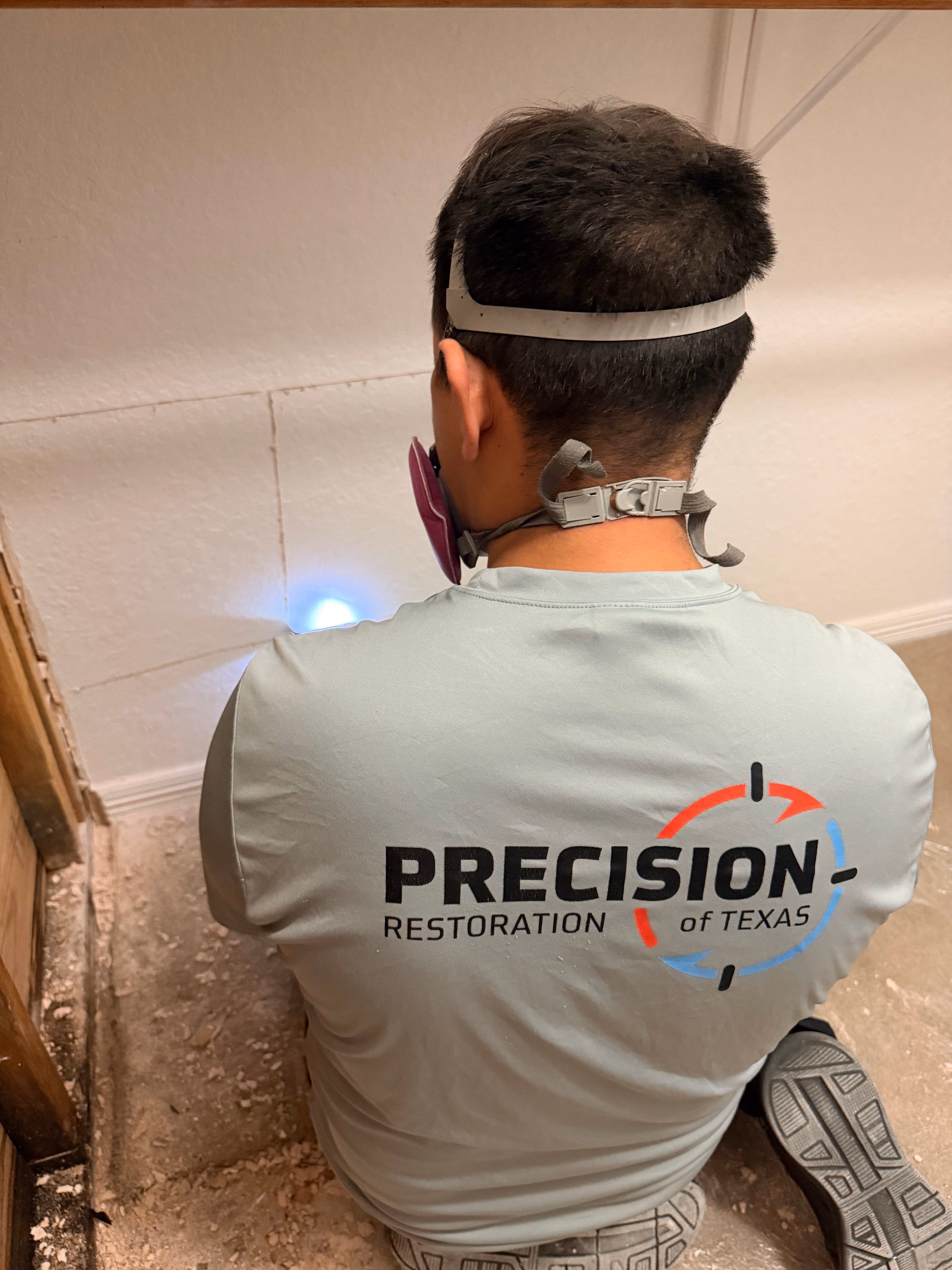 Technician wearing a Precision Restoration of Texas shirt inspecting a wall with a flashlight while kneeling.