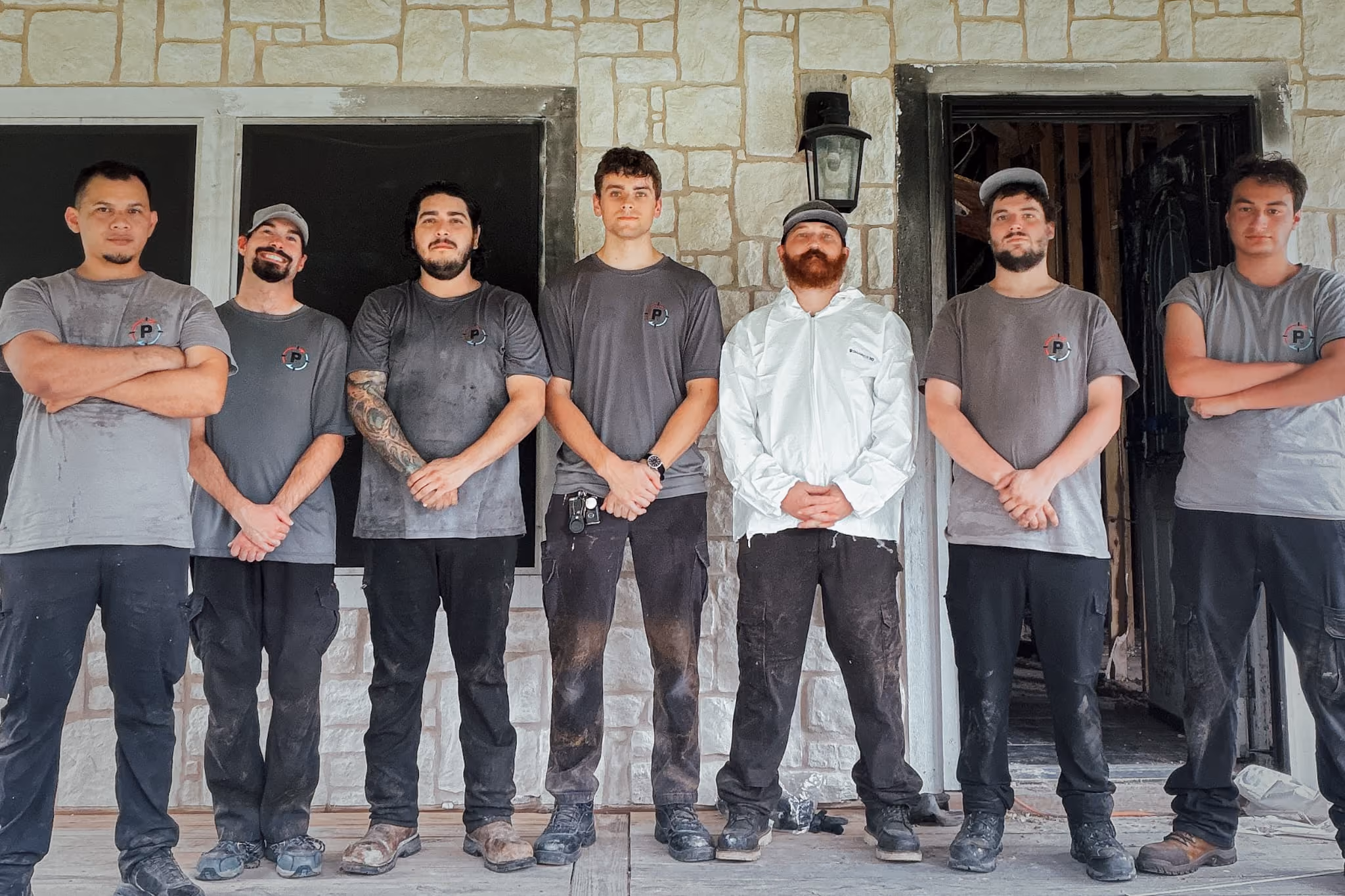 Seven men in work clothes standing side by side outside a stone building with dark windows and a door.