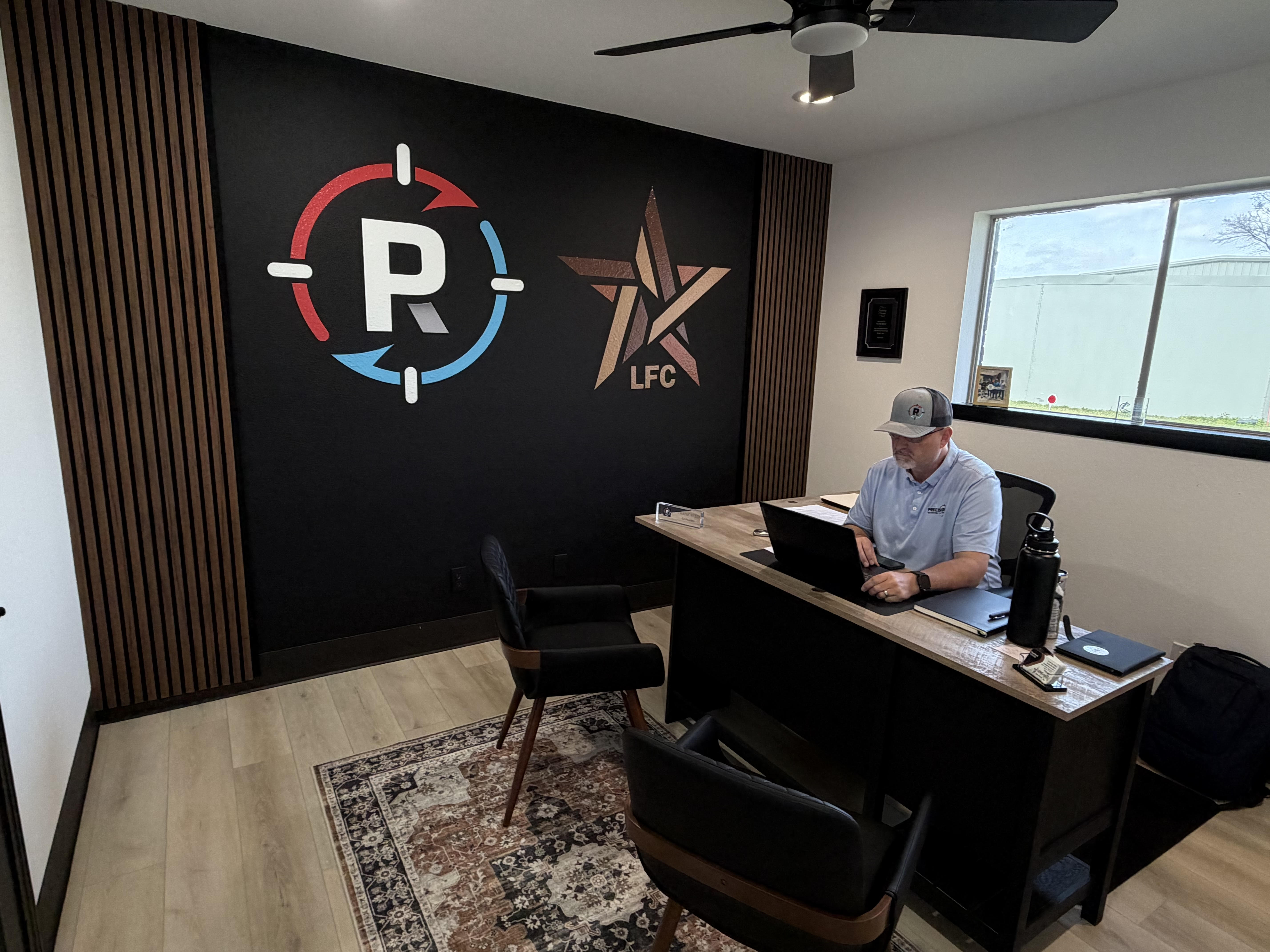 Man wearing cap working on a laptop at a desk in an office with logo wall and window.