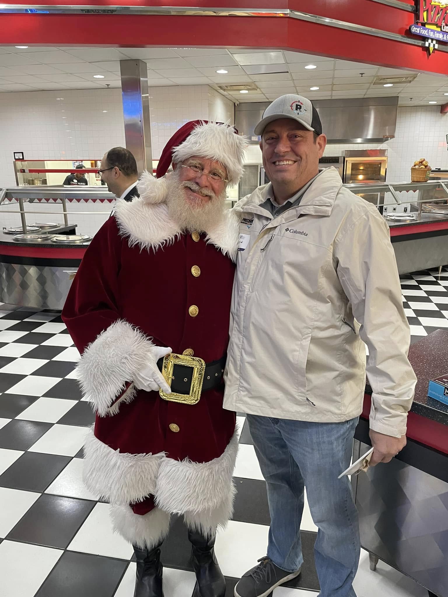 Smiling man in beige jacket standing with person dressed as Santa Claus in restaurant with black-and-white floor.