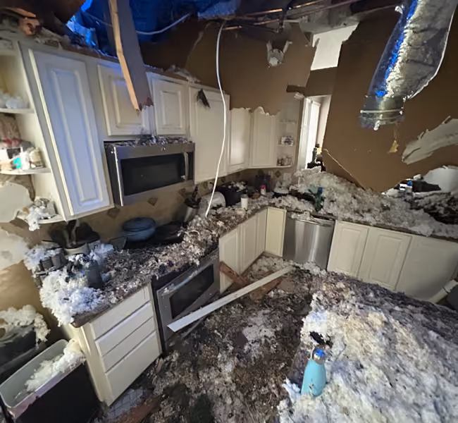Severely damaged kitchen with collapsed ceiling, insulation and debris covering counters and floor.
