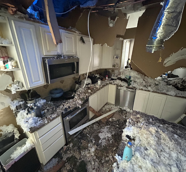 Severely damaged kitchen with collapsed ceiling, insulation and debris covering counters and floor.